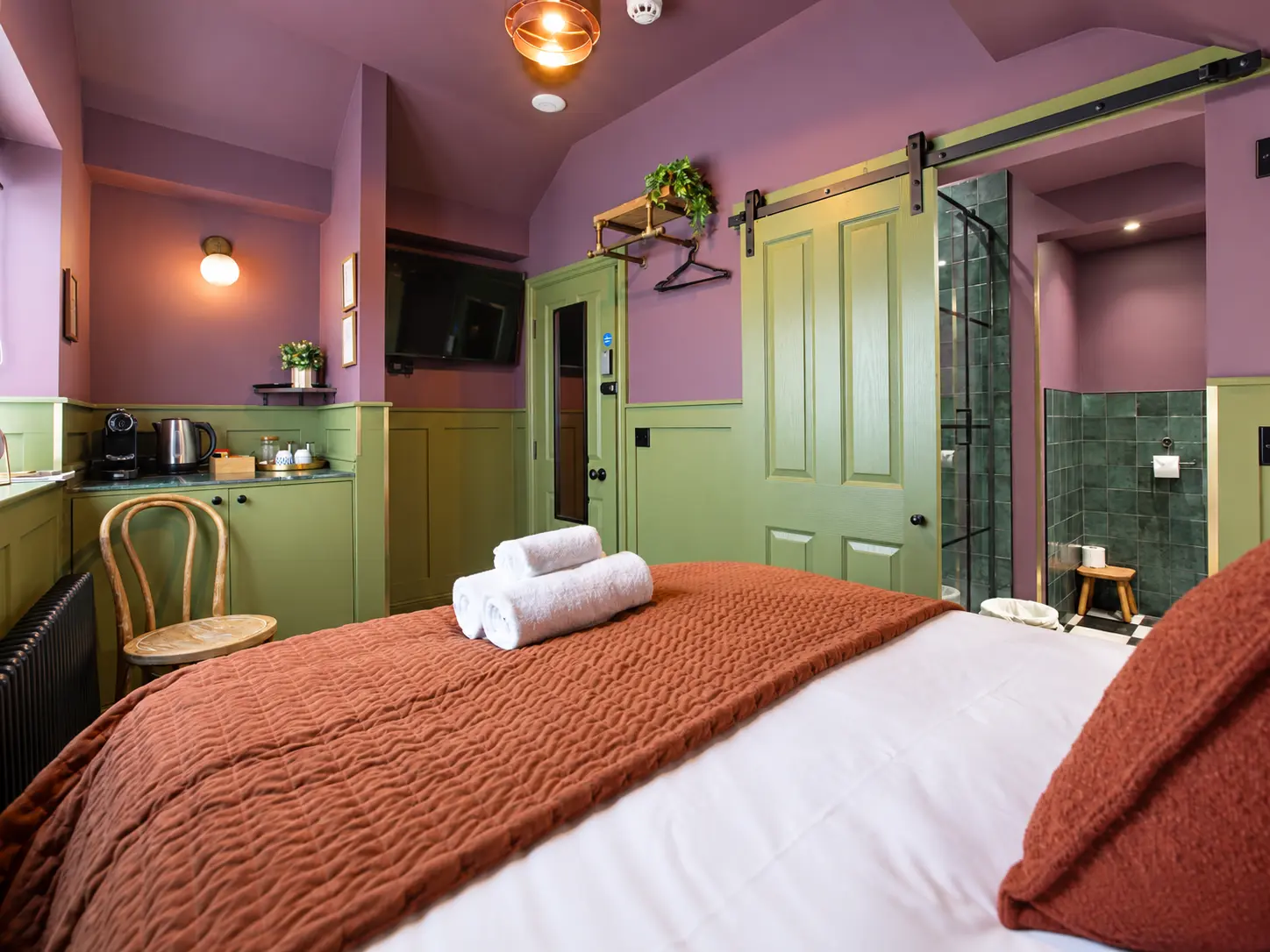 Room 4 at The Linen Rooms — king bed seen toward the coffee station and wall-mounted TV, with the olive barn door open to the ensuite